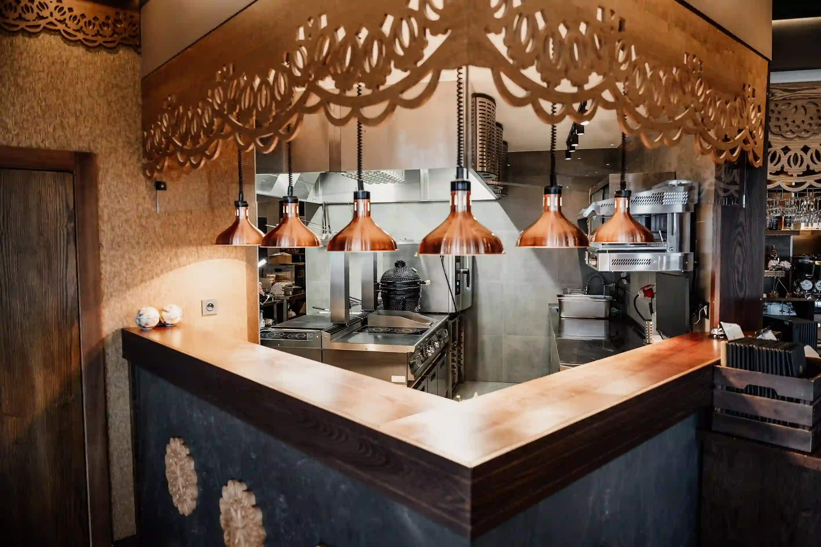 Otwarta kuchnia restauracyjna z ladą ekspozycyjną, miedzianymi lampami grzewczymi i profesjonalnym wyposażeniem gastronomicznym.